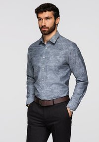 Homme barbu portant une chemise ajustée grise à motifs floraux boutonnée et rentrée dans un pantalon noir avec une ceinture marron, les mains dans les poches.