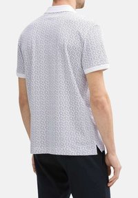 Polo shirt blanc avec un motif géométrique noir, manches courtes avec un liseré blanc et fentes sur les côtés pour plus de confort.