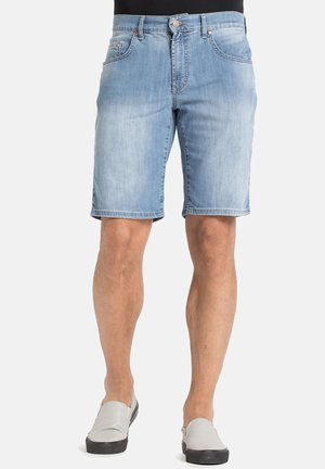 Uomo con pantaloncini di jeans azzurri chiari e scarpe slip-on bianche, in piedi contro uno sfondo chiaro e uniforme.