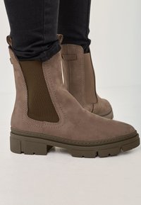 Brown suede ankle boots with elastic side panels, square toe, and chunky rubber sole. Stitched detailing and textured accents visible.