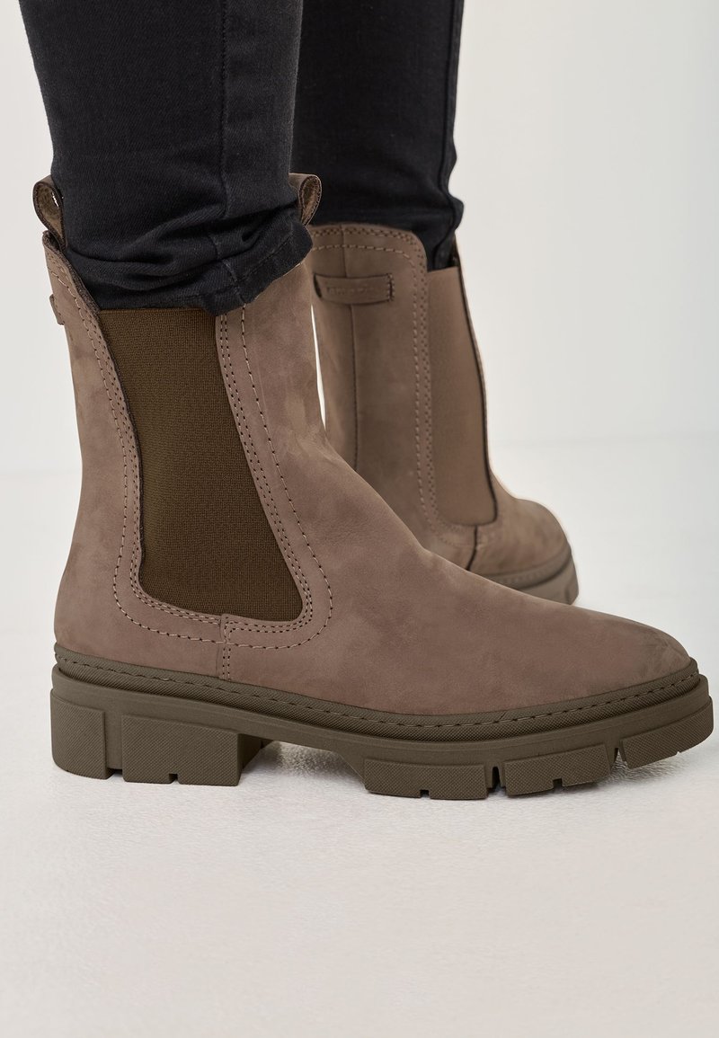 Brown suede ankle boots with elastic side panels, square toe, and chunky rubber sole. Stitched detailing and textured accents visible.