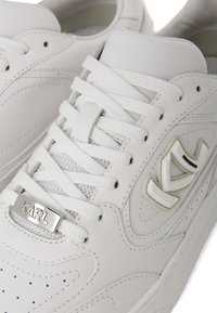 Baltas ādas sporta apavi ar teksturētām acs paneļiem, plakaniem baltiem šņorēm un sudraba logo plāksni. Akcentēti ar uzskatāmu zīmolu un gludām virsmām.