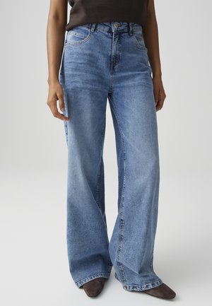 Vrouw die wijde blauwe denim jeans en donkere schoenen draagt, staand tegen een effen lichtgrijze achtergrond.