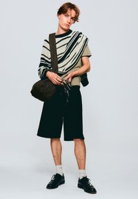 Pantaloni neri abbinati a una maglietta grigia, sovrastati da un pullover a righe. Una borsa a tracolla marrone scuro pende da una spalla. Scarpe nere completano il look.