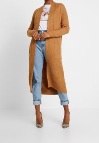 Modell iklädd en lång, beige cardigan över en vit skjorta och blå jeans, kombinerat med klackar med ormskinnsmönster. Enkelt bakgrund.