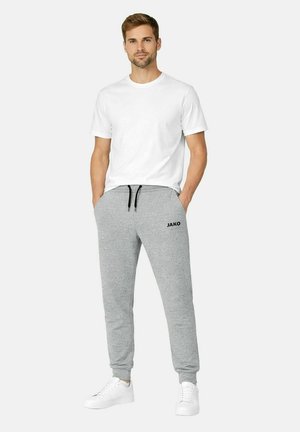 Mann steht mit den Händen in den Taschen, trägt ein weißes T-Shirt, graue JAKO-Sweatpants und weiße Sneaker vor einem einfarbigen hellen Hintergrund.