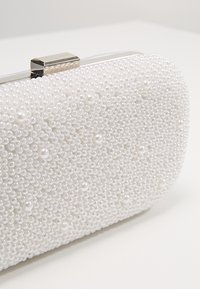 Clutch blanca con adornos de perlas, de forma rectangular redondeada y cierre metálico. Superficie texturizada con perlas blancas de diferentes tamaños.