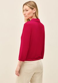 Figurbetonte Bluse mit langen Ärmeln in strahlendem Pink, mit Kragen und gerafftem Saum. Kombiniert mit hellbeigen Hosen aus einem schlichten, strukturierten Stoff.