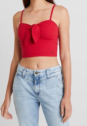 Donna che indossa un top corto rosso con dettaglio nodo frontale e jeans a vita alta blu chiaro su sfondo neutro.