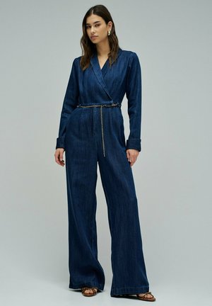 Mujer de pie vistiendo un mono de mezclilla azul oscuro con mangas largas, pantalones anchos, un cinturón delgado de cadena y sandalias marrones con tiras.