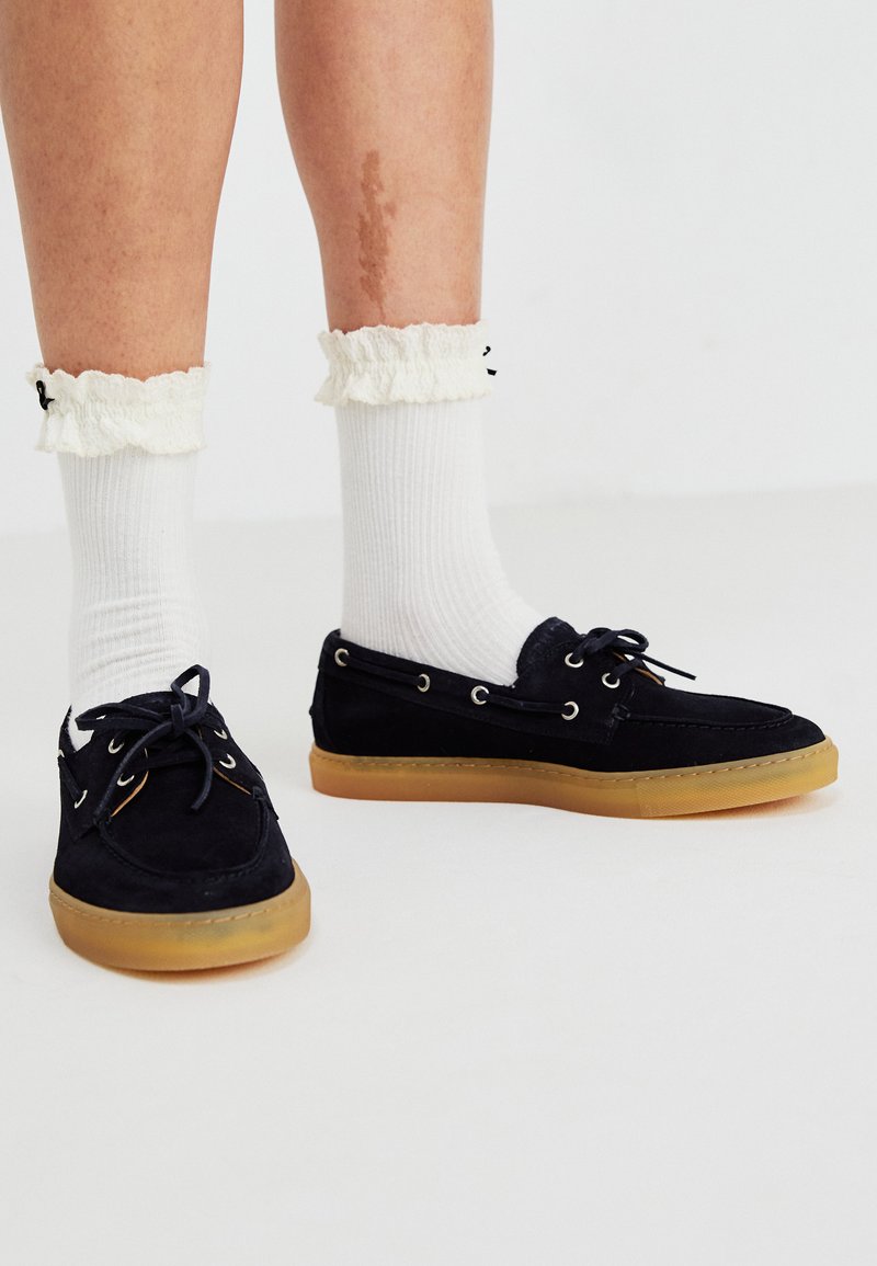 Marineblaue Wildlederschuhe für Boote mit Gummisohlen, ausgestattet mit silbernen Ösen und Schnürsenkeln. Kombiniert mit weißen gerippten Socken und Rüschendetails an den Bündchen.