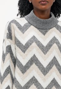 Grauer, beigefarbener und weißer Chevron-Muster-Strickpullover mit engem geripptem Kragen und weichem, strukturiertem Stoff.