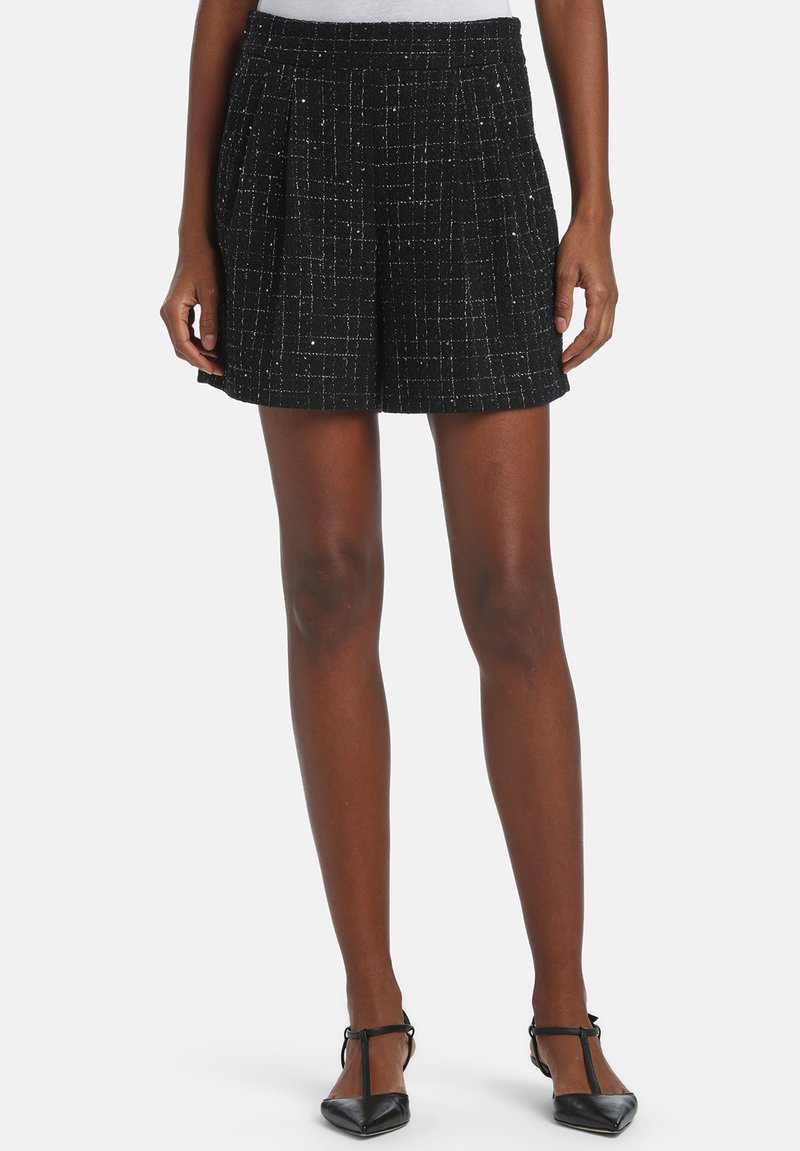 Shorts en tweed noir avec une finition texturée, présentant un motif à petits carreaux subtils et des plis à la taille. Portés avec des chaussures noires à bout pointu.