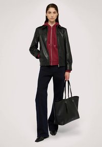 Černá kožená bunda přes bordó mikinu s kapucí, doplněná tmavými džíny a černými botami. Drží velkou černou tašku přes rameno.