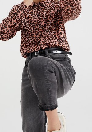 Person iført opkørte sorte jeans, sort bælte, hvide sneakers og en langærmet leopardmønstret skjorte, med det ene knæ løftet.