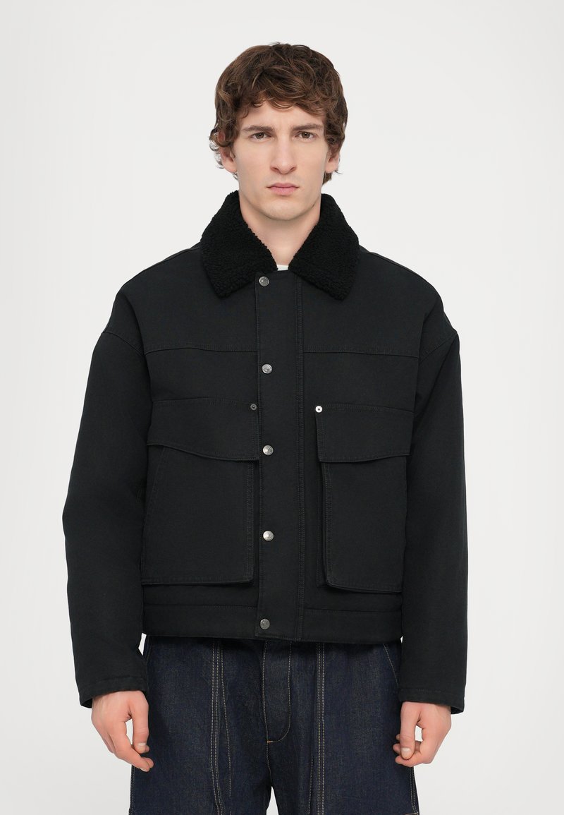 Black denim jacket with a textured black shearling collar, front snap buttons, and two chest pockets. Features a boxy design and wide sleeves.