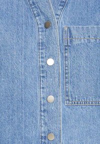 Detailný záber modrého denimového látka so štyrmi kovovými patentkami a štvorcovou náprsné kapsou s viditeľným šitím.