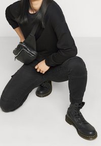 Botas de combate de cuero negro con costuras amarillas, pantalones cargo negros ajustados y una bandolera negra con cremallera. Atuendo texturizado y casual.