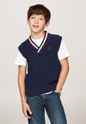 Gilet lavorato a maglia blu navy con scollo a V, caratterizzato da strisce bianche e bordeaux sul colletto, indossato sopra una maglietta bianca e abbinato a jeans di denim.