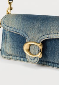 Modra jeans torbica čez ramo z zlato kovinsko zaponko in nastavljivim trakom na belem ozadju.