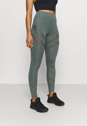 Kobieta w zielonych legginsach z wysokim stanem z panelami z siateczki oraz czarnych butach sportowych, stojąca na tle jednolitego jasnego tła.