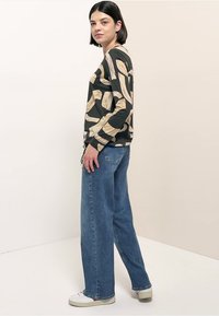Zwart top met lange mouwen en een beige abstract patroon, gecombineerd met lichtblauwe jeans met wijde pijpen. De outfit wordt compleet gemaakt met witte sneakers.