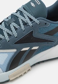 Chaussure de sport présentant un mélange de matériaux bleus et noirs, une tige texturée, des accents en mesh, et un logo blanc avec une semelle en caoutchouc.