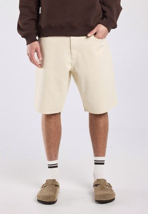 Man draagt beige knielange shorts, witte sokken met zwarte strepen, bruine instapsandalen en een donkerbruine longsleeve.