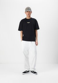 Tricou negru din bumbac cu un mic logo galben, asortat cu pantaloni albi largi și adidași negri. O șapcă gri completează ținuta.
