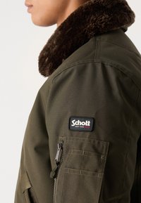 Oliven grøn jakke lavet af holdbart stof, med en brun pelskrave, sidelomme med lynlås og et sort Schott logo-patch på ærmet.