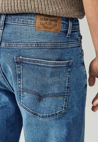 Traperice srednje plave boje, s smešnim kožnim oznakom "DENIM SUPPLY" na pojasu i karakterističnim dizajnom zadnjeg džepa.