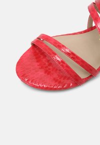 Röda sandaler med skinnande, strukturerade ovandelar och tre tunna remmar över foten. Sulan är platt och slät, med en rundad tådesign.