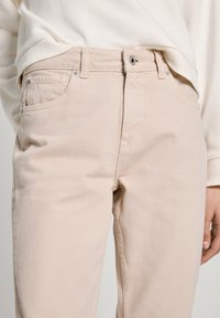 Beige bavlněné džíny se rovnými nohavicemi, s předními kapsami, zapínáním na zip a jemnými švy. Hladká textura a minimální kovové detaily.