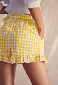 Gelbe Gingham-Shorts mit Rüschensaum, verziert mit weißen Margeriten, die auf den Stoff gestickt sind. Leichtes Baumwollmaterial. Mit Taschen.