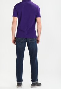 Camisa de polo morada con un collar corto, utilizada con jeans azul oscuro. Los jeans tienen un corte recto y los puños enrollados. Unos zapatos grises completan el conjunto.