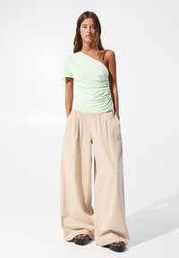 Mintgroene top met één schouder en gerimpelde details, gecombineerd met ruime beige broeken van lichtgewicht stof en hogere zwarte plateau schoenen.