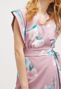 Robe portefeuille rose avec des motifs floraux en bleu et blanc. Caractérisée par des manches larges et une taille ceinturée. Tissu lisse, léger et fluide.