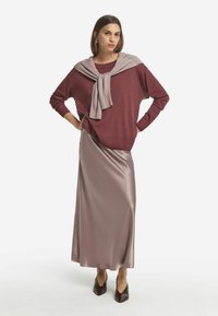 Bordeauxfarbener Pullover, leicht grau darunter, am Hals gebunden, kombiniert mit einem langen, glatten, satinähnlichen, hellen Taupe-Rock und dunklen spitzen Schuhen.