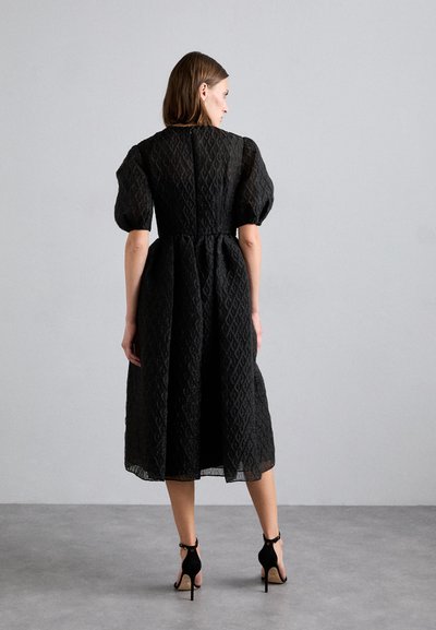 Black midi dress with puff sleeves, textured fabric featuring a diamond pattern, cinched waist, and a back zipper. Paired with black heels.