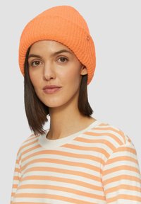 Pomarańczowa, prążkowana czapka beanie o miękkiej teksturze, z małym metalowym akcentem, w zestawieniu z kremową koszulą w paski.