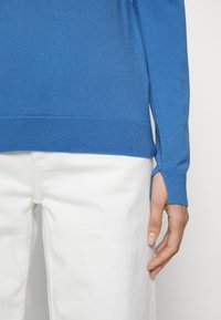 Pull tricot bleu avec fentes latérales aux poignets, associé à un pantalon en denim blanc. Texture lisse et coupe ajustée mettent en valeur la forme du vêtement.