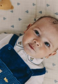 Kék kordbársony baba overall, hímzett medve mintával, fehér galléros inggel, amelyen egy illeszkedő medve díszítés található.
