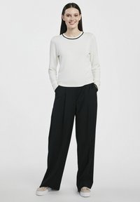 Kremowy sweter z długim rękawem i czarnym dekoltem, w zestawieniu z czarnymi spodniami z wysokim stanem i szerokimi nogawkami oraz beżowymi sneakersami, ręce w kieszeniach.