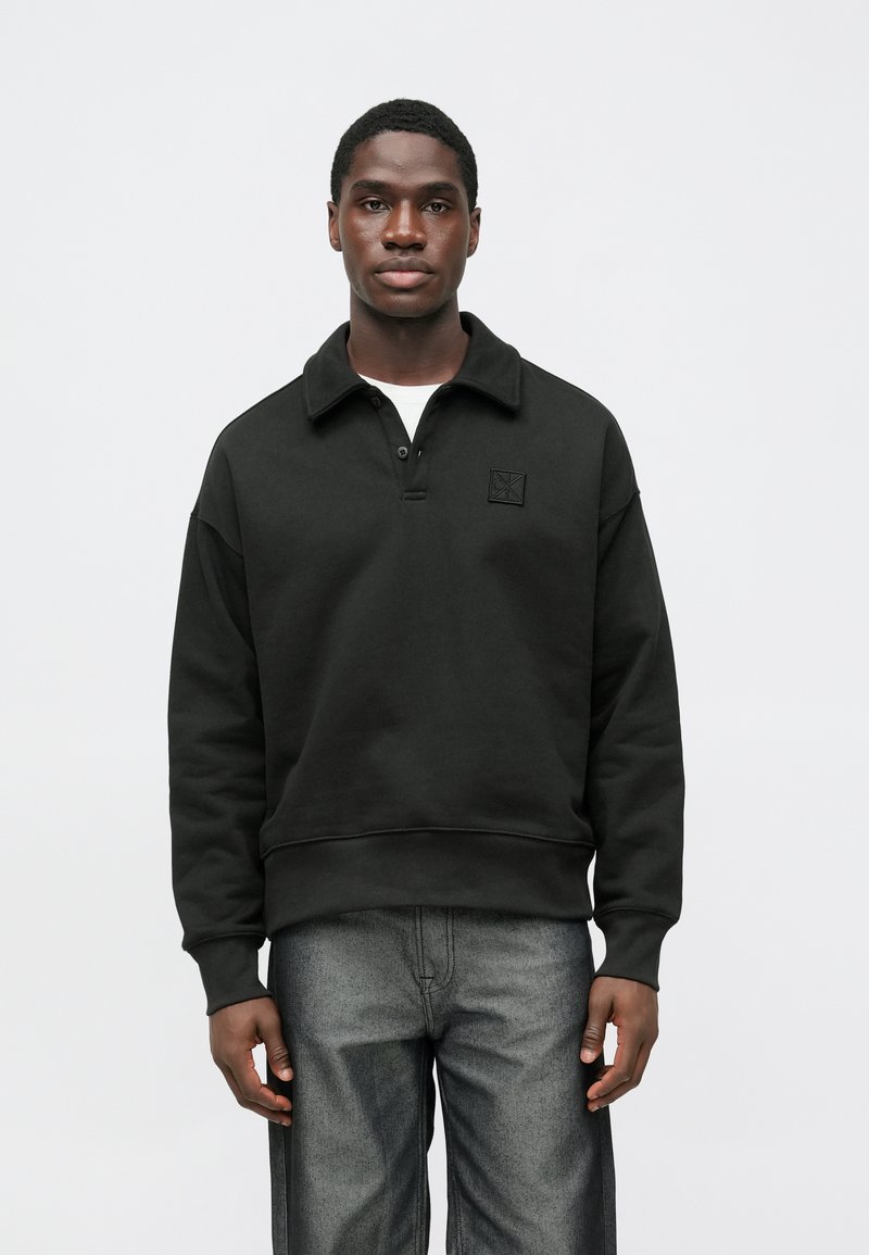 Czarna bluza z kołnierzykiem, dwoma guzikami i teksturowanym logo na piersi. Luźny krój i ściągacze przy rękawach. Nosi się ją na białą koszulę.