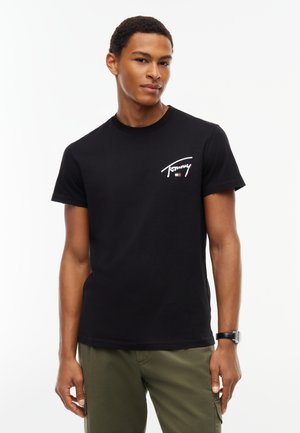 Młody mężczyzna w czarnej koszulce Tommy Hilfiger i oliwkowych spodniach stoi na białym tle.