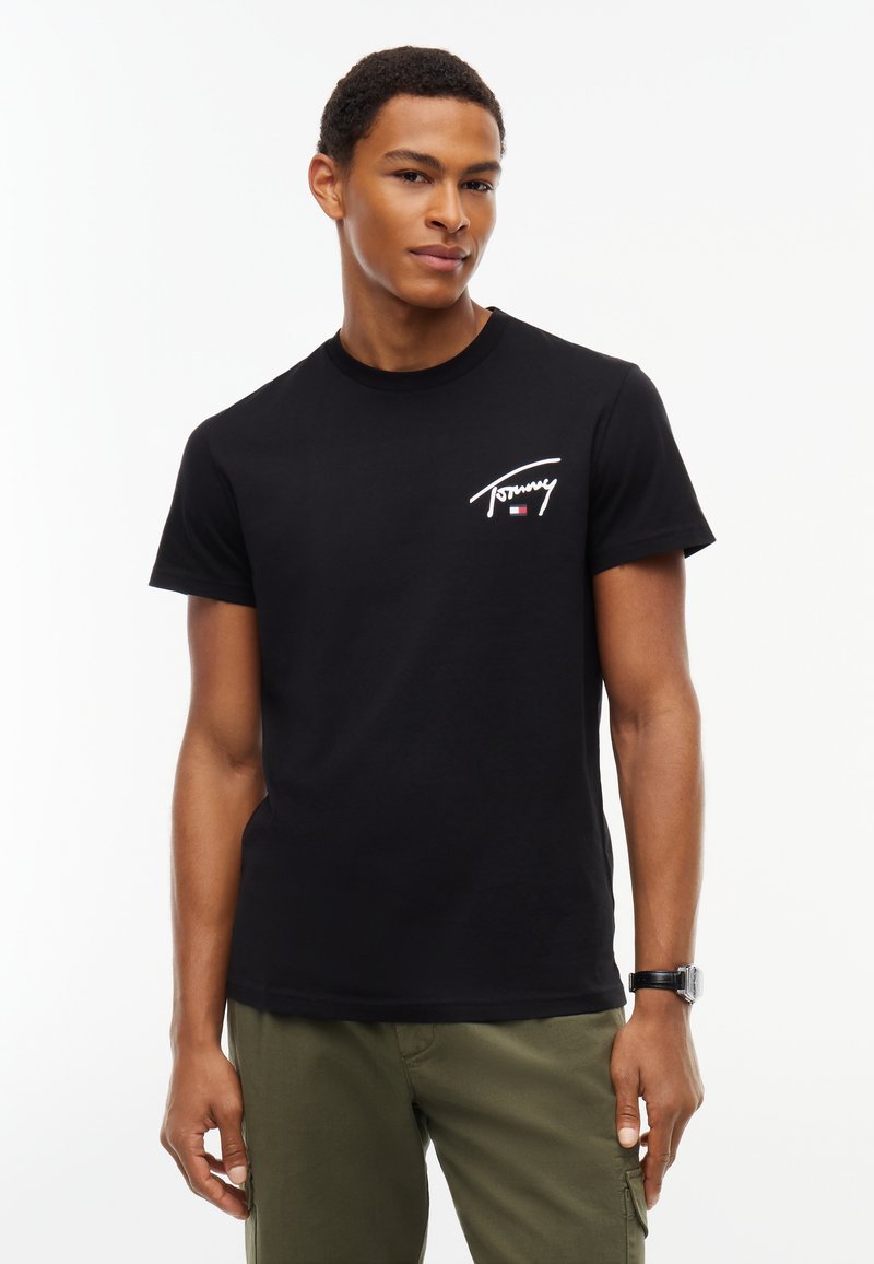 Νέος άντρας που φοράει μαύρο μπλουζάκι Tommy Hilfiger και χακί παντελόνι στέκεται μπροστά σε λευκό φόντο.