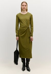 Langärmliges, olivgrünes Kleid mit Knoten-Detail an der Taille, kombiniert mit glänzenden schwarzen kniehohen Stiefeln und einer schwarzen Tote-Tasche.