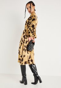 Γυναίκα σε καφέ και μαύρο κα patterned midi φόρεμα, κρατώντας μια τσάντα μαύρη με καρφιά, φορώντας μαύρες μπότες ως το γόνατο με τακούνι, ποζάροντας πλευρικά.