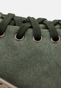 Sneaker verde din suede cu ochiuri metalice rotunde, șireturi plate și o suprafață texturată. Prezintă cusături pe margini și o talpă bej.