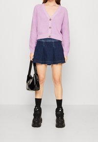 Ljuslila stickad cardigan med tre knappar, kombinerad med en mörk denimfållad kjol. Svart lackhandväska och kraftiga svarta sneakers.